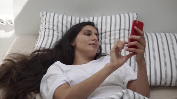Awakened young woman is typing messages on her smartphone, lying in bed - Záběry, video
