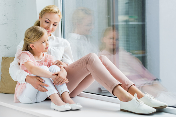 beautiful happy mother and daughter sitting together on windowsill - Photo, Image