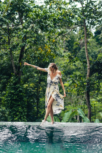 attractive blond woman in dress walking near swimming pool with green plants on background, ubud, bali, indonesia - Photo, Image