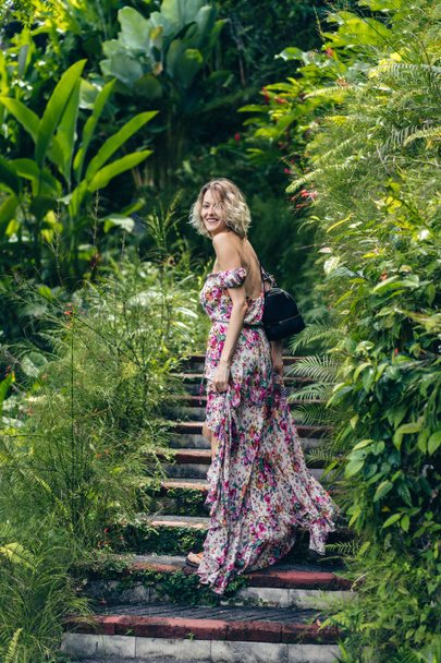 Seitenansicht einer lächelnden blonden Frau im Kleid, die auf Stufen inmitten grüner Pflanzen steht, ubud, bali, indonesien - Foto, Bild