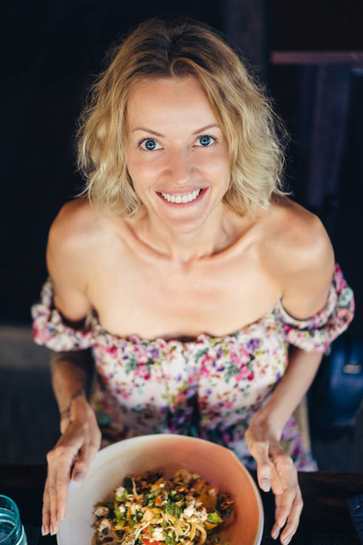 hoge hoekmening van vrolijke blonde vrouw zittend aan tafel met schotel en camera kijken - Foto, afbeelding
