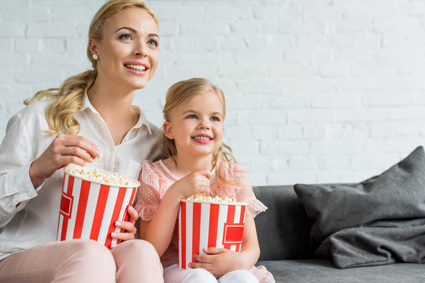 happy mother and daughter eating popcorn and looking away at home  - Fotó, kép