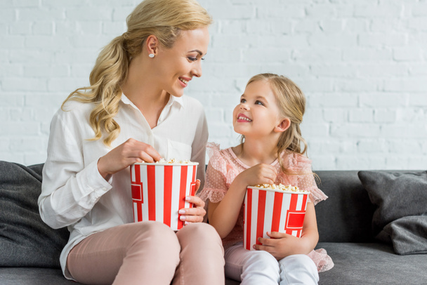happy mother and daughter smiling each other while eating popcorn at home - Photo, Image
