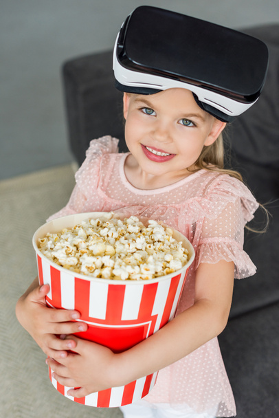 beautiful child in virtual reality headset holding box with popcorn and smiling at camera - Fotoğraf, Görsel