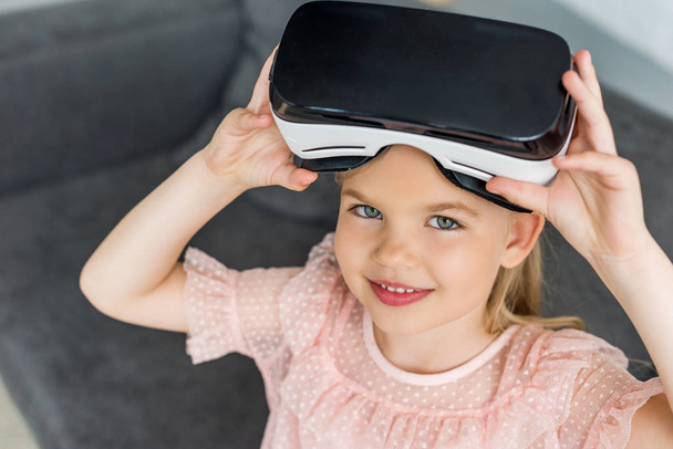 hoge hoekmening van schattig kind in virtual reality headset glimlachend op camera thuis - Foto, afbeelding
