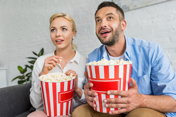 excited couple eating popcorn and looking away at home - Photo, Image