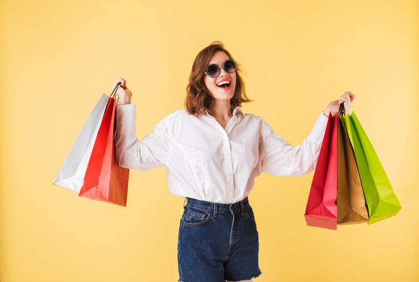 Portrait of young joyful lady in sunglasses standing with colorful shopping bags in hands on over pink background. Happy woman standing in white shirt and denim shorts  - Fotoğraf, Görsel