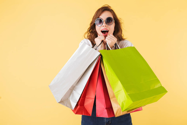 Portrait of young surprised lady in sunglasses standing with colorful shopping bags in hands and looking in camera on over pink background - 写真・画像