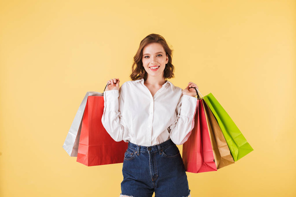 Portrait of beautiful smiling lady standing with colorful shopping bags in hands and joyfully looking in camera on over pink background. Pretty woman standing in white shirt and denim shorts - Photo, Image