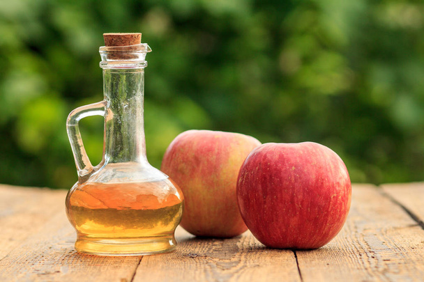Apple vinegar in glass bottle with cork and fresh red apples on wooden boards with blurred green natural background. Organic food for health - Photo, Image