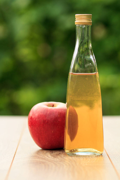 Appel azijn in glazen fles met metalen dop en verse rode appel op houten planken met groene natuurlijke achtergrond. Biologische voeding voor gezondheid - Foto, afbeelding