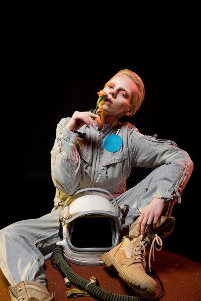 attraktive Astronautin im Raumanzug mit Helm auf dem Planeten sitzend und Rosenblume in der Hand - Foto, Bild