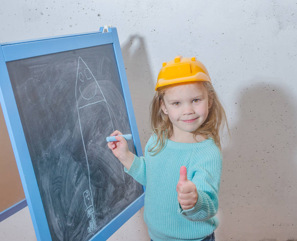child engineer, designer, girl in a helmet paint a house on a black chalk Board - Photo, Image