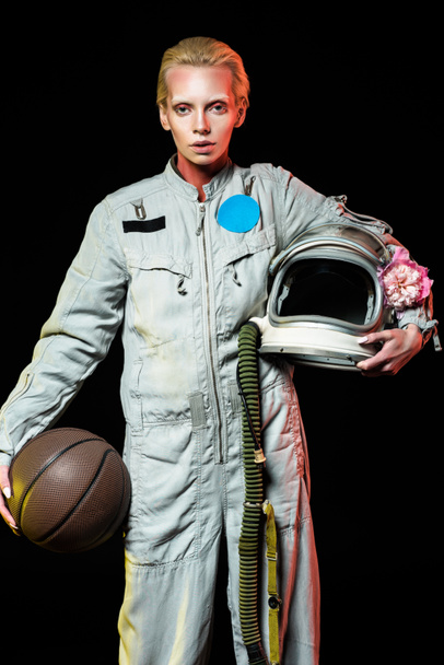 attractive female astronaut in spacesuit holding basketball and helmet with flower, isolated on black - Photo, Image