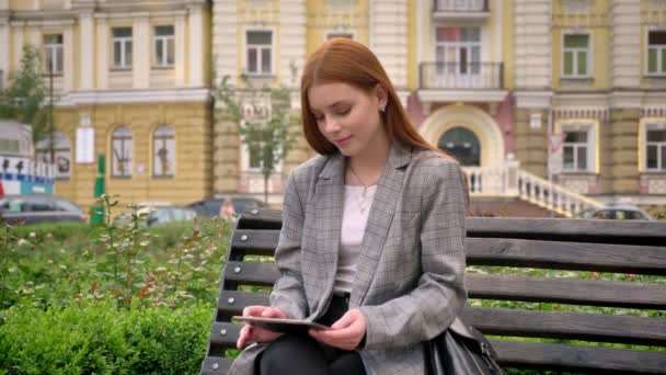Young beautiful ginger woman in jacket sitting on bench and holding tablet, city street background - 映像、動画