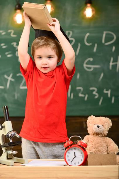 Child, pupil on cheerful face near microscope. Wunderkind concept. First former interested in studying, education. Kid boy in graduate cap holds book in classroom, chalkboard on background - Foto, Imagen
