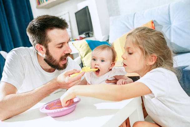 Good looking young man eating breakfast and feeding her baby girl at home - Photo, image