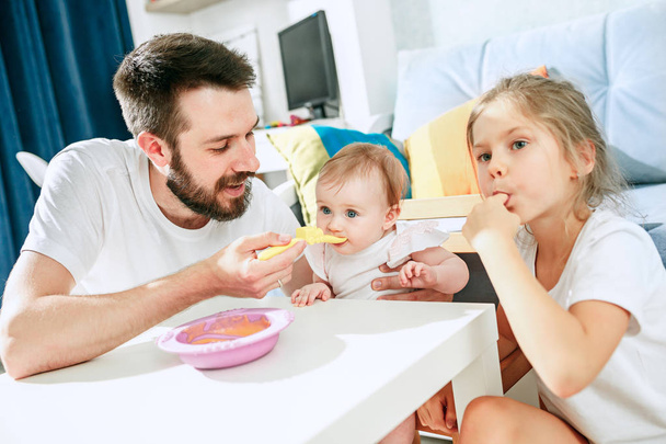 Good looking young man eating breakfast and feeding her baby girl at home - Zdjęcie, obraz
