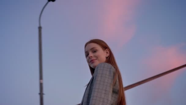 Young beautiful woman with long ginger hair looking in camera and turning around, blue sky above, circling around view, low angle - Footage, Video