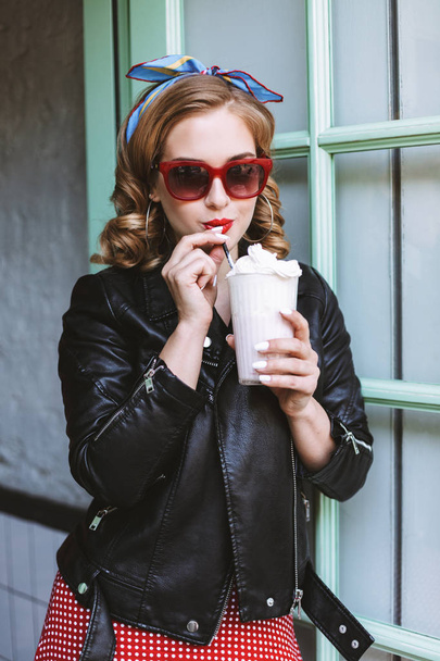 Красуня в сонцезахисних окулярах і шкіряній куртці стоїть біля дверей в кафе і п'є молочний коктейль
  - Фото, зображення