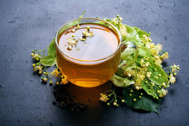 Bylinný čaj s květy lime ve skleněném pohárku na tmavý podklad. Užitečné a chutný nápoj. Prostředky pro tradiční medicíně k léčbě nachlazení. Boční pohled - Fotografie, Obrázek