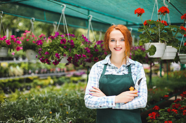 Прекрасний усміхнений флорист в фартусі стоїть з садовими ножицями в руці. Молода леді радісно дивиться в камеру під час роботи з квітами
 - Фото, зображення
