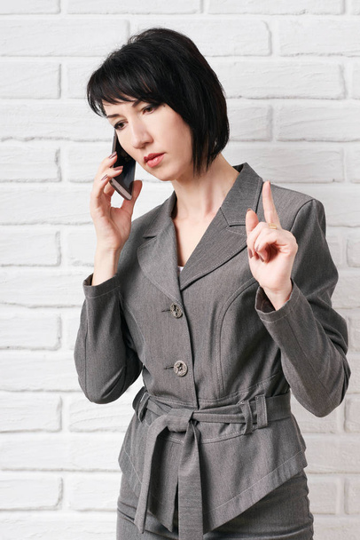 business woman dressed in a gray suit talking on the phone, stay in front of a white wall - Fotoğraf, Görsel