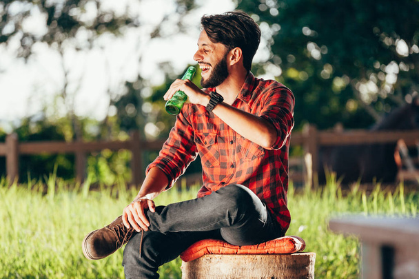 Young farmer sitting on bench and drinking beer with pasture and corral in background - Fotoğraf, Görsel