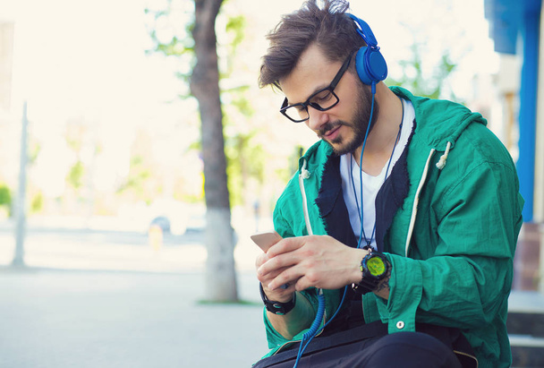 Young trendy man listening to music with headphones and smartphone while sitting on street - Photo, image