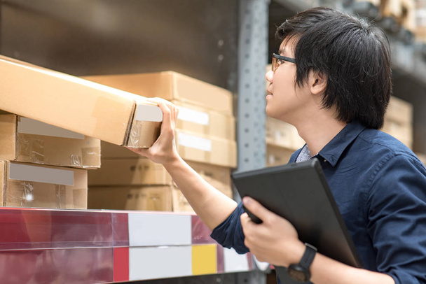 Young Asian man doing stocktaking of product in cardboard box on shelves in warehouse by using digital tablet. physical inventory count concept - Photo, Image
