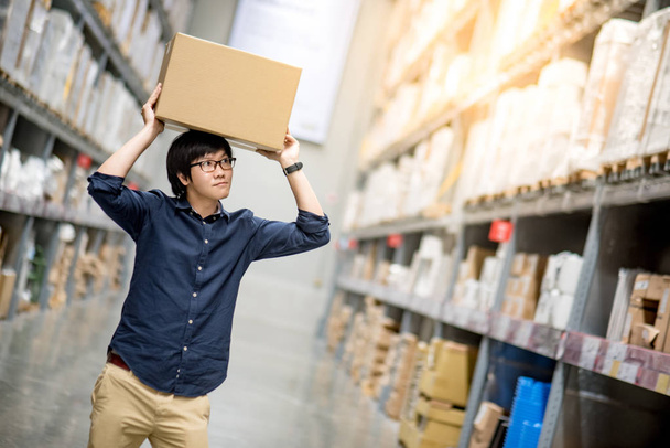 Young Asian man carrying cardboard box over head between row of shelves in warehouse, shopping warehousing or working pick and packing concepts - Photo, Image