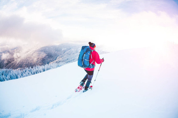 Een man in sneeuwschoenen en stokken van de trekking in de bergen. Winter reis. Klimmen voor een klimmer tegen een mooie hemel met wolken. Actieve levensstijl. Beklimming van de berg door de sneeuw. - Foto, afbeelding
