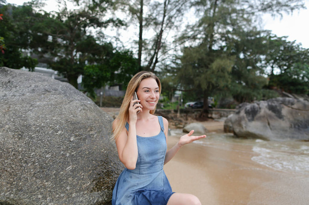 Молодая женщина, сидящая на песке у моря и камней, разговаривает по смартфону
. - Фото, изображение