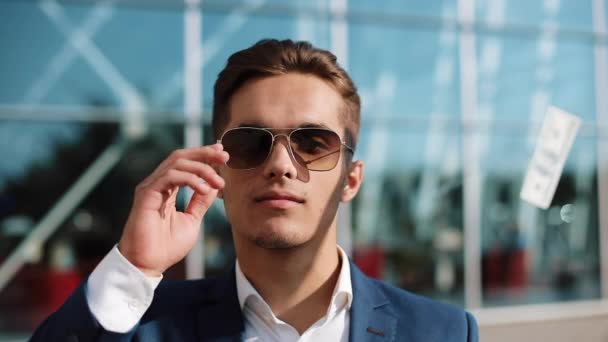 Portrait of a young businessman with sunglasses in focus looking at camera while money flying down all over the plac. Money rain on the businessman in slow motion. - Filmagem, Vídeo