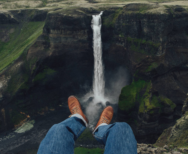 travel and Hiking to Iceland's highest waterfall . hiker  photographing his legs from above. A man is sitting on the edge of a cliff - Valokuva, kuva