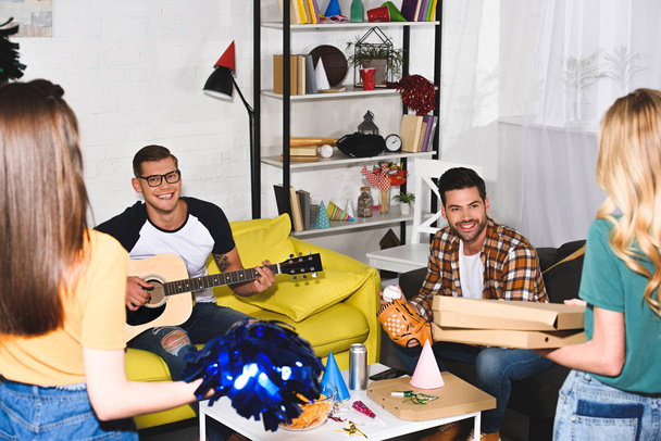 smiling young men playing guitar and looking at girls holding pizza at home party - Foto, Bild