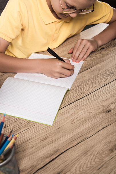partial view of schoolboy in eyeglasses writing in empty textbook at table with colour pencils - Photo, Image