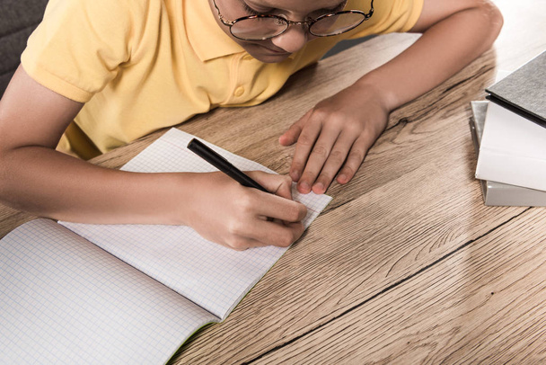 vista parziale di scolaro in occhiali che scrive in libro di testo vuoto a tavola con pila di libri
 - Foto, immagini