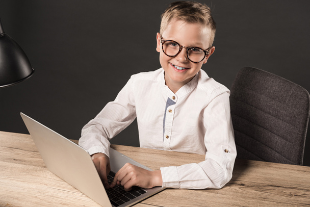happy little boy in eyeglasses looking at camera while sitting at table with laptop and lamp on grey background  - Zdjęcie, obraz