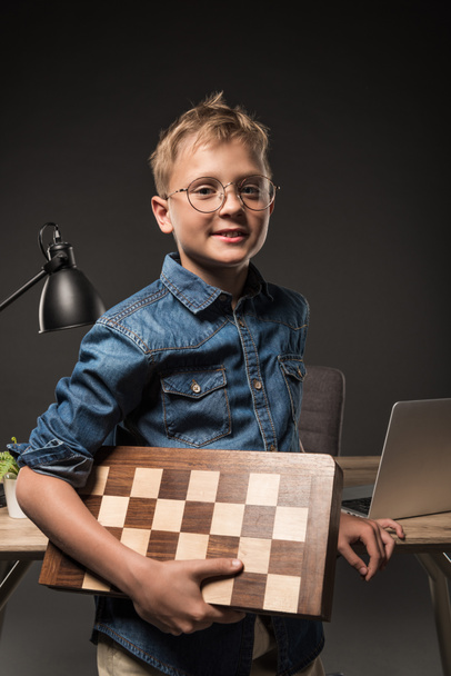 smiling little boy in eyeglasses holding chessboard near table with laptop and lamp on grey background  - Foto, Imagem