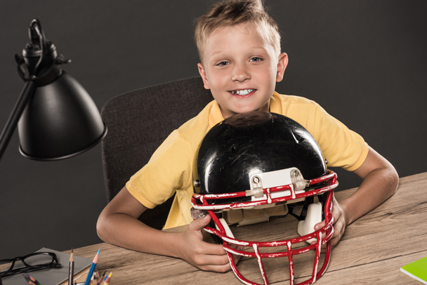 happy schoolboy sitting with american football helmet at table with eyeglasses, lamp, colour pencils and books on grey background  - Fotó, kép