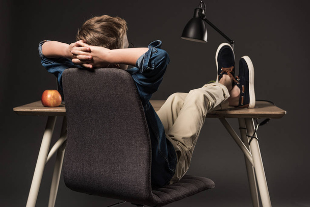 rear view of schoolboy resting with legs on table on grey background  - Photo, Image