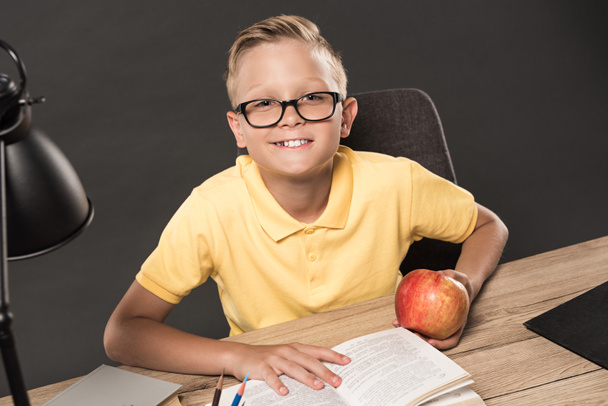 smiling schoolboy in eyeglasses looking at camera while sitting at table with apple, books and lamp on grey background  - 写真・画像