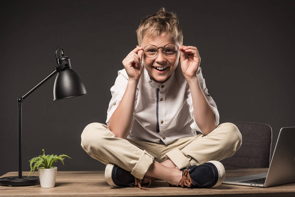 smiling little boy in eyeglasses sitting on table with plant, lamp and laptop on grey background   - Fotoğraf, Görsel
