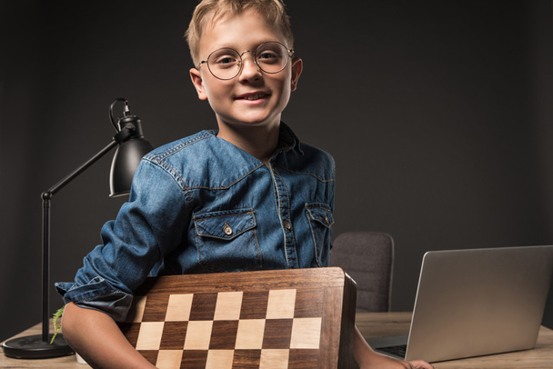 niño feliz en anteojos mirando a la cámara y sosteniendo tablero de ajedrez cerca de la mesa con el ordenador portátil y la lámpara sobre fondo gris
  - Foto, imagen