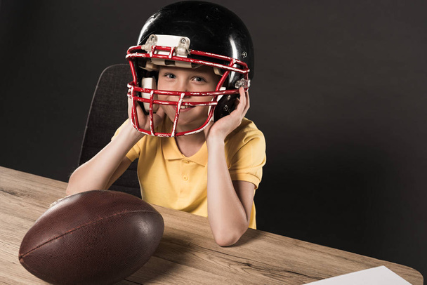 usměvavý chlapeček v ochranné přilbě u stolu s míčem na americký fotbal na šedém pozadí  - Fotografie, Obrázek