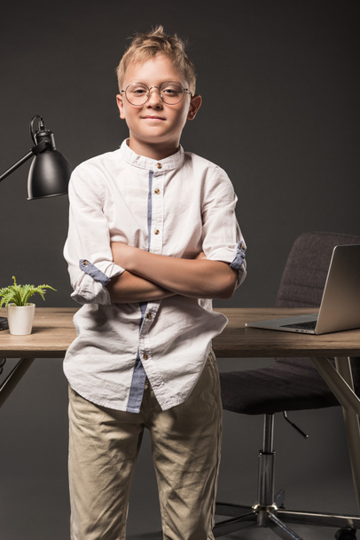 bambino in occhiali con le mani incrociate in piedi vicino al tavolo con pianta, computer portatile e lampada su sfondo grigio
  - Foto, immagini