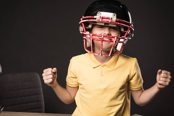 smiling little boy in protective helmet for american football gesturing by hands on grey background  - Фото, изображение