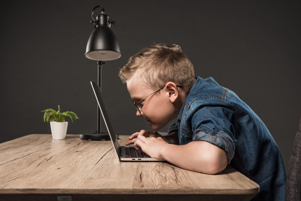 Seitenansicht eines kleinen Jungen in Brille mit Laptop am Tisch mit Lampe und Pflanze auf grauem Hintergrund  - Foto, Bild
