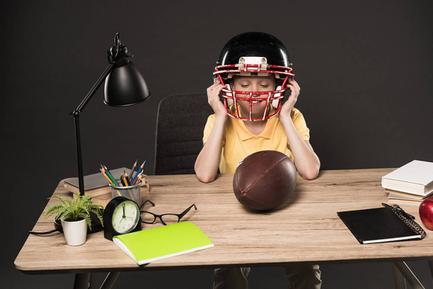 schooljongen Amerikaans voetbal helm op te zetten en zittend met benen op tafel met bal, boeken, plant, lamp, kleur potloden, apple, klok en leerboek over grijze achtergrond  - Foto, afbeelding